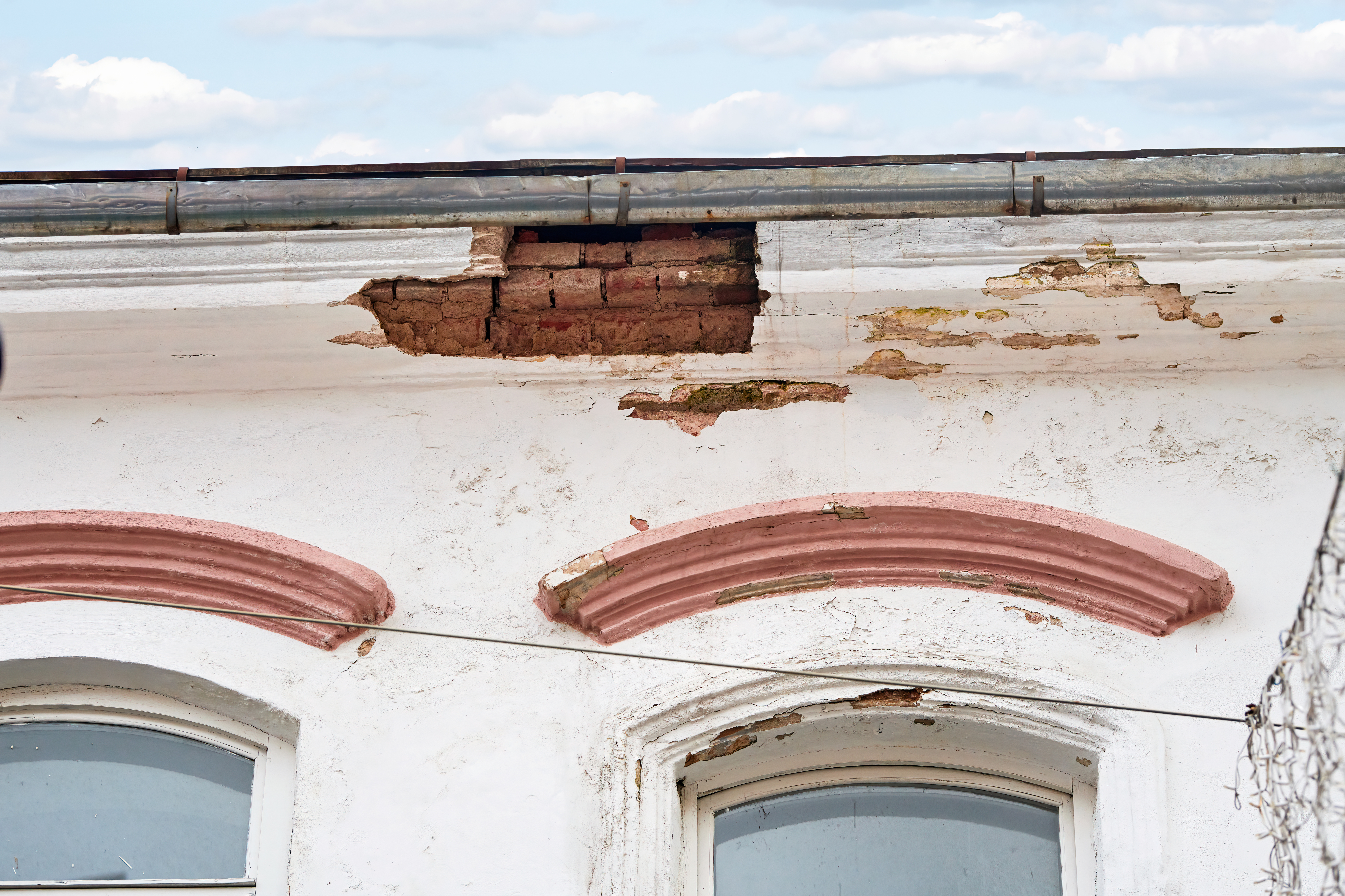 Decaying historic building with visible wall damage, chipped paint, crumbling roof edges, architectural wear, weathering, lack of preservation efforts. Damaged facade of an aging building with cracks